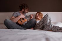 Веселий молодий чоловік з дредлоками і жінка з рудим волоссям сидить на ліжку і розважається з закритими очима, граючи на укулелевій гітарі, проводячи час разом вдома на вихідних — стокове фото