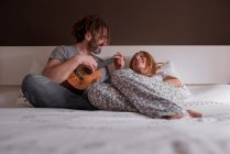 Jovem alegre com dreadlocks e mulher com cabelo vermelho sentado na cama e se divertindo com os olhos fechados tocando guitarra ukulele enquanto passam o tempo juntos em casa no fim de semana — Fotografia de Stock