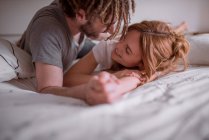 Homem macio com dreadlocks abraçando e beijando mulher com cabelo vermelho enquanto deitado no estômago na cama juntos e relaxante no fim de semana — Fotografia de Stock