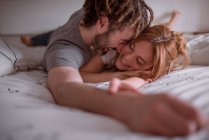 Tender man with dreadlocks hugging and kissing woman with red hair while lying on stomach on bed together and relaxing on weekend — Stock Photo