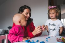Positive Mutter in lässiger roter Kleidung sitzt mit Kindern am Tisch und spielt mit Knetmasse, während sie Zeit zu Hause verbringt — Stockfoto