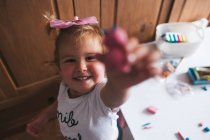 Улыбающаяся маленькая девочка в белой повседневной рубашке, держа в руке плазитциновую игрушку ручной работы и дотрагиваясь до камеры, играя дома — стоковое фото