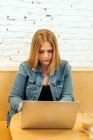 Mujer autónoma concentrada en ropa casual sentada a la mesa en un espacio de trabajo luminoso y escribiendo en netbook durante el trabajo remoto - foto de stock