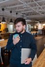 Cheerful male freelancer in wireless earphones and with laptop leaning on table in creative workspace and enjoying hot beverage while having break during remote work — Stock Photo