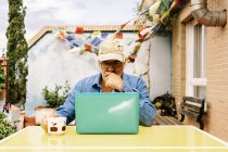 Удумливий чоловік похилого віку в шапці і джинсовій сорочці зосереджується на екрані з інтересом, сидячи за столом з чашкою смачного напою і використовуючи нетбук у дворі заміського будинку — стокове фото