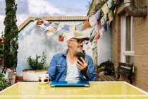 Задумчивый старший мужчина в кепке и просматривает смартфон, сидя за ноутбуком с кружкой вкусного горячего напитка на заднем дворе загородного дома в солнечный день — стоковое фото
