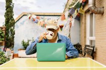 Thoughtful elderly male in cap and denim shirt focusing on screen with interest while sitting at table with mug of tasty beverage and using netbook in yard of country house — Stock Photo