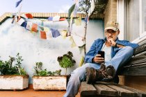 Спокойный задумчивый мужчина в очках и кепке, фокусирующийся на экране и сообщениях с помощью смартфона, расслабляясь на деревянной скамейке на заднем дворе коттеджа в пригороде — стоковое фото