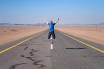 Homem alegre em roupas casuais desfrutando de liberdade e aventura durante a viagem enquanto salta em estrada vazia no vale do deserto com montanhas no horizonte — Fotografia de Stock