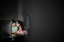 Feliz joven madre con su pequeño hijo y su hija sosteniendo el teléfono inteligente mientras están sentados juntos en la mesa con la computadora y viendo videos divertidos en la habitación oscura en casa - foto de stock