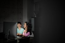 Щаслива молода мати з маленьким сином і дочкою тримає смартфон, сидячи разом за столом з комп'ютером і дивлячись смішне відео в темній кімнаті вдома — стокове фото