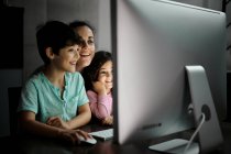 Счастливая молодая мать с маленьким сыном и дочерью держат смартфон, сидя вместе за столом с компьютером и смотреть смешное видео в темной комнате дома — стоковое фото
