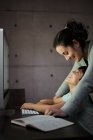 Вид сбоку молодой женщины, объясняющей учебное задание положительному сыну, сидящему за столом с компьютером и учебником во время онлайн-урока дома — стоковое фото
