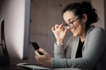 Side view of serious concentrated young female in casual wear and eyeglasses sitting at table with computer with white blank screen and browsing smartphone while working remotely at evening time at home — Stock Photo