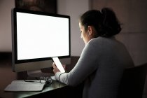Seitenansicht einer ernsten, konzentrierten jungen Frau in Freizeitkleidung und Brille, die am Tisch mit Computer mit weißem leeren Bildschirm sitzt und im Smartphone surft, während sie abends fern zu Hause arbeitet. — Stockfoto