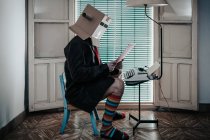 Человек в картонной коробке и полосатых носках сидит на стуле с ретро-пишущей машинкой и читающей бумагой — стоковое фото