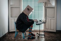 Чоловік у картонній коробці та смугастих шкарпетках сидить на стільці, друкуючи на ретро друкарській машинці — стокове фото