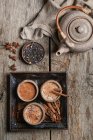 Vista superior de Masala chai servido en cuencos de cerámica con anís estrellado y palos de canela dispuestos en mesa de madera con tetera y trozo de tela - foto de stock