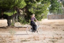 Спокойная женщина в летнем наряде прогуливается с велосипедом в парке в солнечный день и смотрит в сторону — стоковое фото