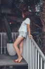Low angle side view of happy slim female in gray casual shirt and shorts with smartphone touching sunglasses and looking away with interest while leaning on railing at wooden staircase in comfortable resort hotel on Bali — Stock Photo