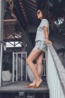 Baixo ângulo vista lateral de feliz fêmea slim em cinza camisa casual e shorts com smartphone tocando óculos de sol e olhando para longe com interesse, enquanto se inclina sobre trilhos em escada de madeira em confortável hotel resort em Bali — Fotografia de Stock