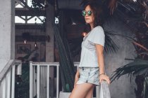 Низкий угол зрения на счастливую стройную женщину в серой повседневной рубашке и шортах со смартфоном касаются солнцезащитных очков и с интересом смотрят в сторону, опираясь на перила на деревянной лестнице в комфортабельном курортном отеле на Бали — стоковое фото