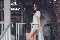 Vista lateral de ángulo bajo de mujer delgada feliz en camisa casual gris y pantalones cortos con smartphone tocando gafas de sol y mirando hacia otro lado con interés mientras se apoya en la barandilla en la escalera de madera en el cómodo hotel resort en Bali - foto de stock