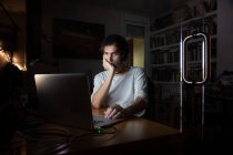 Concentrated young male remote specialist in casual wear talking on mobile phone and using laptop while working on project in dark room at home during evening time — Stock Photo