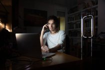 Konzentrierter junger männlicher Fernspezialist in Freizeitkleidung, der mit Handy und Laptop spricht, während er abends im dunklen Raum zu Hause an einem Projekt arbeitet — Stockfoto