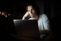 Konzentrierter junger männlicher Fernspezialist in Freizeitkleidung, der mit Handy und Laptop spricht, während er abends im dunklen Raum zu Hause an einem Projekt arbeitet — Stockfoto