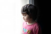 Vista lateral de la niña triste de pie cerca de la ventana y mirando hacia fuera mientras pasa tiempo en casa - foto de stock