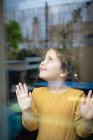 Спокійна маленька дівчинка з кучерявим волоссям, що стоїть біля вікна і задумливо дивиться, проводячи час вдома і мріючи про пригоди — стокове фото