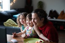 Веселая молодая женщина с милыми маленькими дочерьми сидит за столом и наслаждается видео-разговор с друзьями через ноутбук, проводя вечер дома — стоковое фото