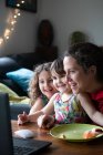 Весела молода жінка з милою маленькою донькою сидить за столом і насолоджується відео розмовою з друзями через ноутбук, проводячи вечір вдома — стокове фото