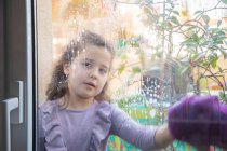 Позитивна маленька дівчинка в повсякденному одязі з пляшкою розпилювача миючого скла в кімнаті з веселкою, намальованою на вікні під час коронавірусної карантини — стокове фото
