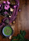 Зверху керамічна чаша свіжого зеленого смузі з гарбузовим насінням і кунжутом в композиції з фіолетовою ніжною тканиною і букетом зі свіжих фіолетових ромашкових квітів у вазі, розміщеній на коричневому дерев'яному столі — стокове фото