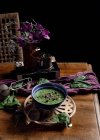 From above of ceramic bowl of fresh green smoothie with pumpkin seeds and sesame in composition with purple delicate fabric and bouquet of fresh violet Daisy flowers in vase placed on brown wooden table — Stock Photo