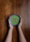 Вид зверху на невизначену жінку, що тримається обома руками керамічна маленька чаша свіжого зеленого смузі з додаванням гарбузового насіння та кунжуту на коричневий дерев'яний стіл — стокове фото
