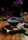 Von oben Keramikschüssel mit frischem grünem Smoothie mit Kürbiskernen und Sesam in Komposition mit lila zartem Stoff und Bouquet frischer violetter Gänseblümchen in Vase auf braunem Holztisch — Stockfoto