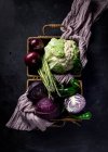 Вид зверху на різні свіжі стиглі овочі з фіолетовим рушником у плетеному кошику, розміщеному на темній поверхні як концепція здорової їжі — стокове фото