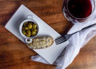 Вид сверху на чашу с маринованными оливками и брускеттой на тарелке, поставленной на стол с бокалом красного вина на кухне — стоковое фото