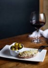 Top view of bowl with pickled olives and bruschetta on plate placed on table with glass of red wine in kitchen — Stock Photo