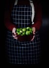 Жіночий кухар у пробному фартусі стоїть з мискою свіжої Брюссельської капусти на чорному тлі на кухні. — стокове фото