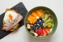 Сверху вкусная миска для завтрака с нарезанными фруктами и ягодами на столе возле тоста с жареным яйцом и ветчиной — стоковое фото