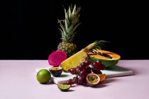 Bodegón con frutas tropicales: papaya en rodajas, piña, pitaya y uvas sobre tabla de cortar de mármol - foto de stock