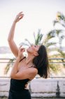 Витончена жіноча танцівниця в момент виконання елемента з витягнутими руками і закритими очима на літній терасі на фоні пальмових дерев — стокове фото