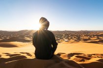Обратный вид на неузнаваемого туриста, сидящего на песчаной дюне и любующегося величественными пейзажами заката в пустыне в Марокко — стоковое фото