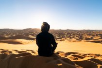 Vista traseira do turista irreconhecível sentado na duna de areia e admirando majestoso cenário do pôr do sol no deserto em Marrocos — Fotografia de Stock