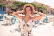 Alluring adult female in straw hat wearing light dress looking at camera while standing alone on cafe terrace against blue and white furniture in sunlight — Stock Photo