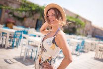 Alluring adult female in straw hat wearing light dress looking at camera while standing alone on cafe terrace against blue and white furniture in sunlight — Stock Photo